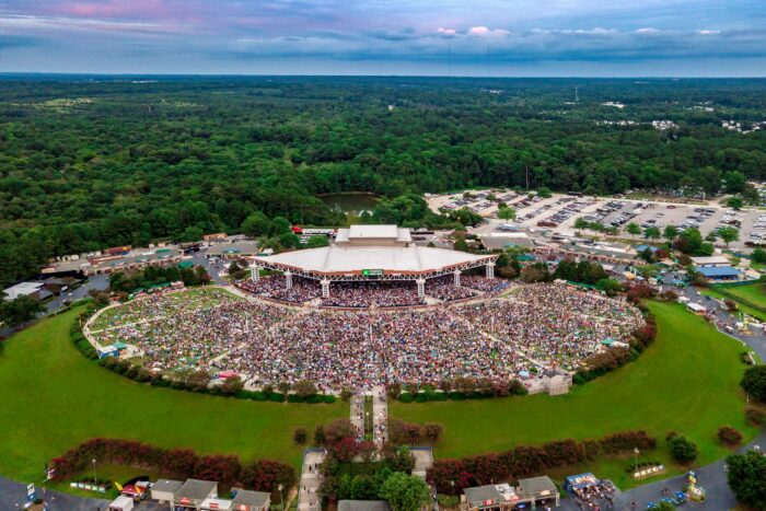 Aerial view of a packed concert at Coastal Credit Union Music Park in Raleigh, North Carolina, surrounded by wooded landscapes and outdoor lawn seating.